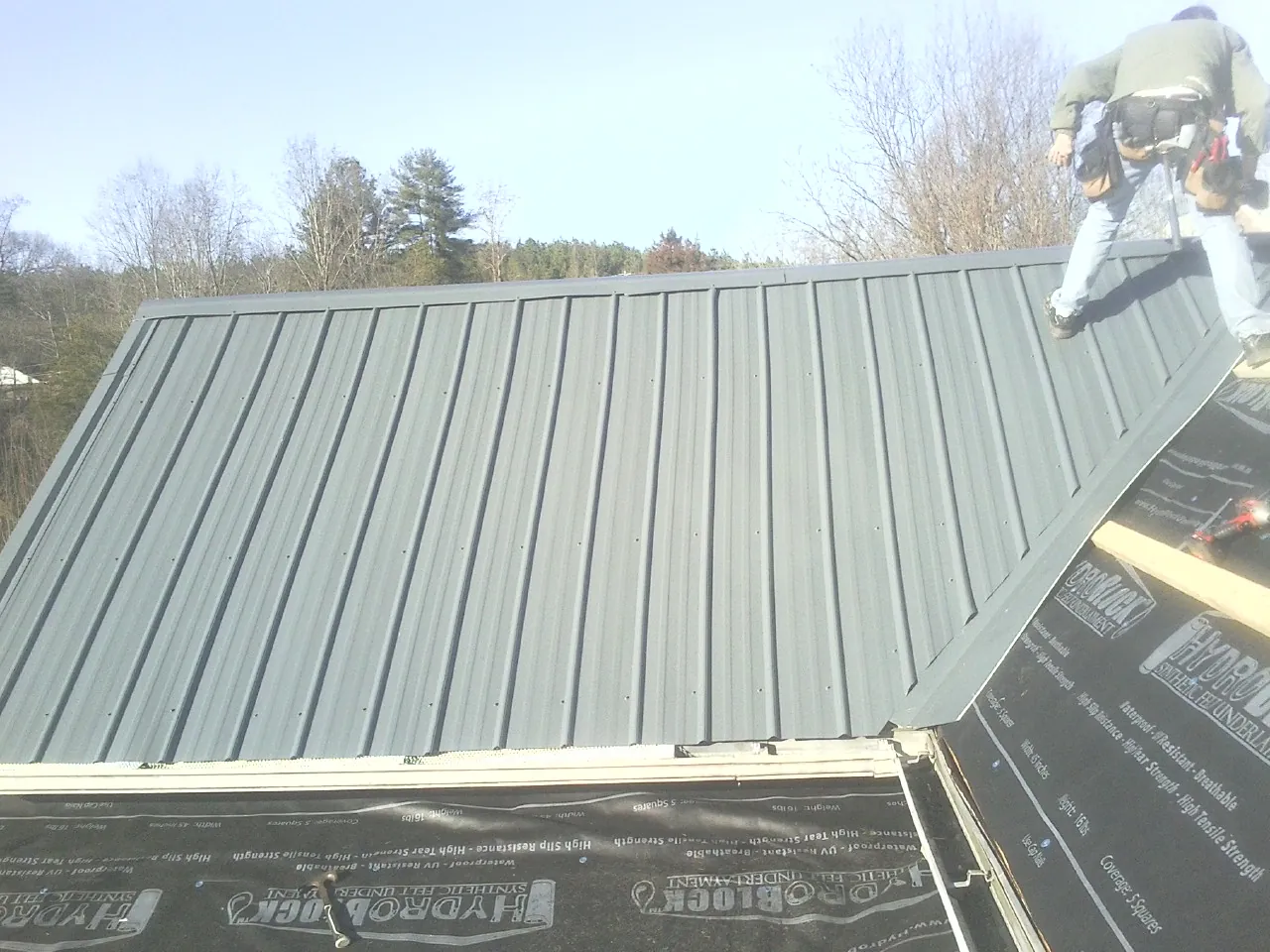 Crew installing dark green standing seam panels over synthetic underlayment in Temperanceville