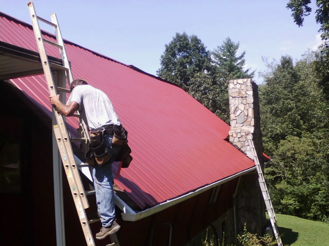 Roofer on a ladder beside a red metal roof and stone chimney — Soffit & Fascia Repair by Roofing Brothers in Temperanceville